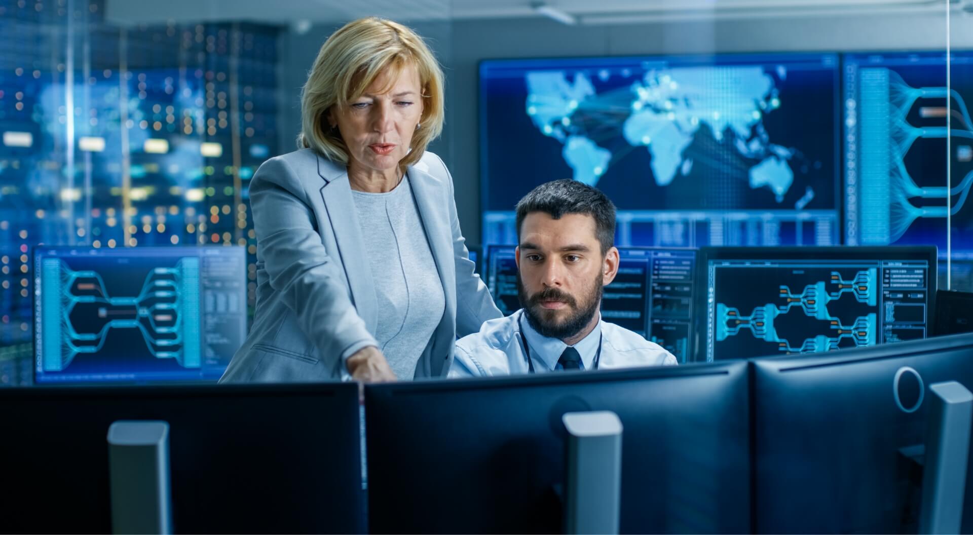 woman providing direction to male counterpart behind computer monitors in command center environment 
